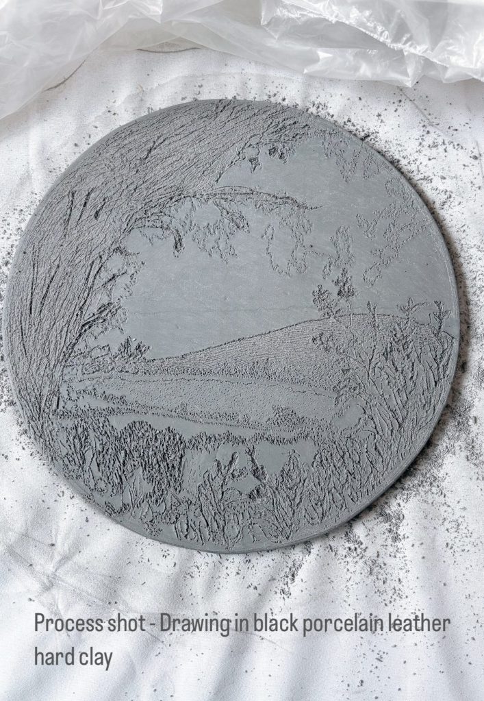 Process shot of a round piece of black porcelain leather hard clay with a detailed landscape drawing, surrounded by clay dust on a white cloth.