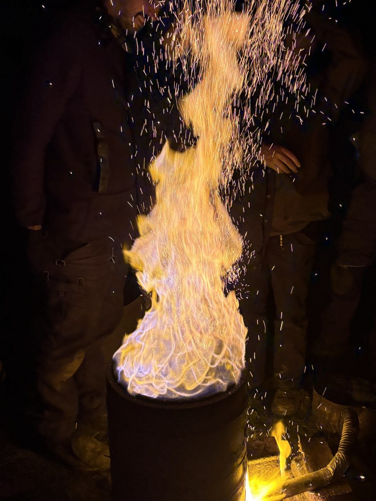 A flaming fire emerging from a barrel with sparks flying in the dark, highlighting the surrounding figures.